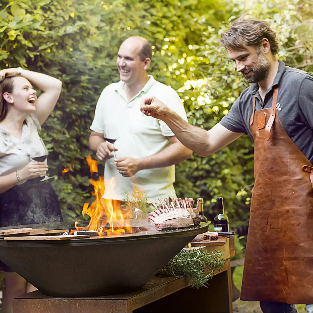 A man grilling a rack of lamb on a large fire pit during an outdoor gathering, with two people enjoying drinks and laughing in the background amidst lush greenery