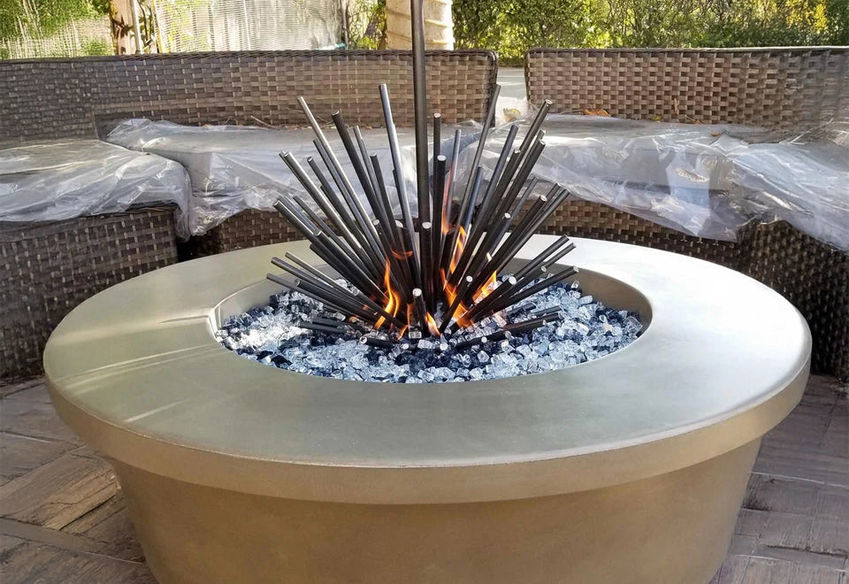 Contemporary round fire pit with a striking spiky metal sculpture centerpiece, surrounded by reflective fire glass, adding an artistic touch to an outdoor seating area with wicker furniture.