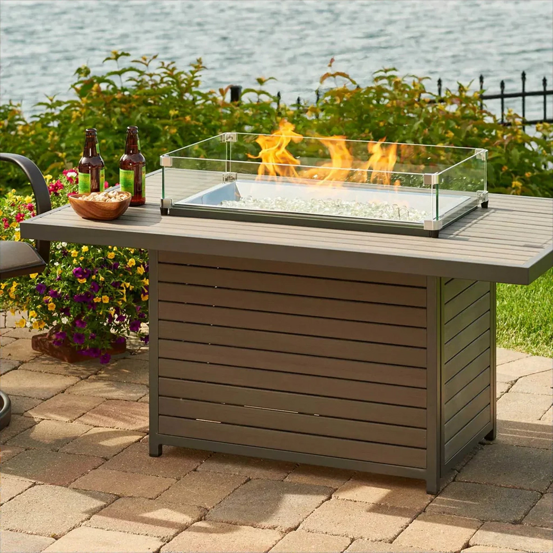 Rectangular fire pit table with a glass wind guard on a patio, featuring two bottles of beer and a bowl of snacks, overlooking lush greenery and a peaceful waterfront setting