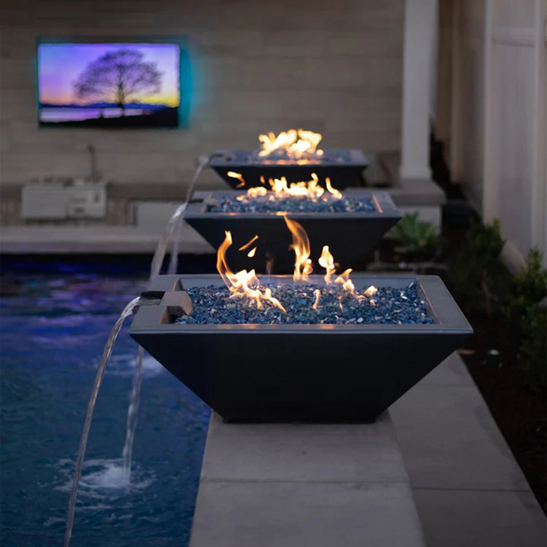 Three fire and water bowls with blue fire glass and flowing water lined up along a swimming pool, creating a stunning nighttime ambiance. A television displaying a serene landscape is visible in the background