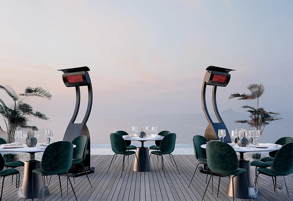 Modern freestanding patio heaters warming a stylish outdoor dining area with green velvet chairs and an ocean view at sunset.