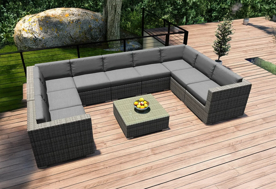 Spacious gray wicker outdoor sectional sofa with plush gray cushions, arranged on a wooden deck with natural greenery and a stone feature in the background.