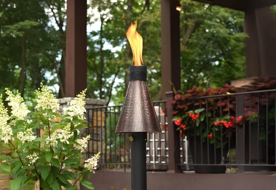 Elegant hammered copper patio lighting torch with an open flame, adding warmth and ambiance to an outdoor garden setting with lush greenery and floral accents.