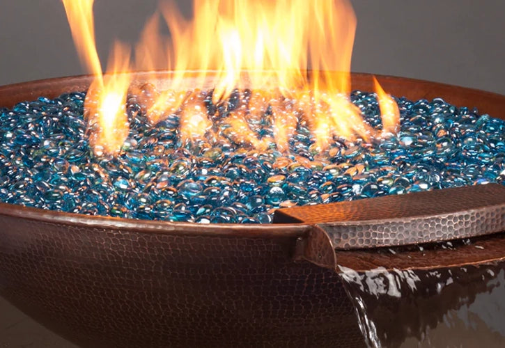 Close-up view of a hammered metal fire pit filled with blue fire glass, featuring flames and a waterfall effect, where water flows over the edge, creating a dynamic and serene atmosphere.