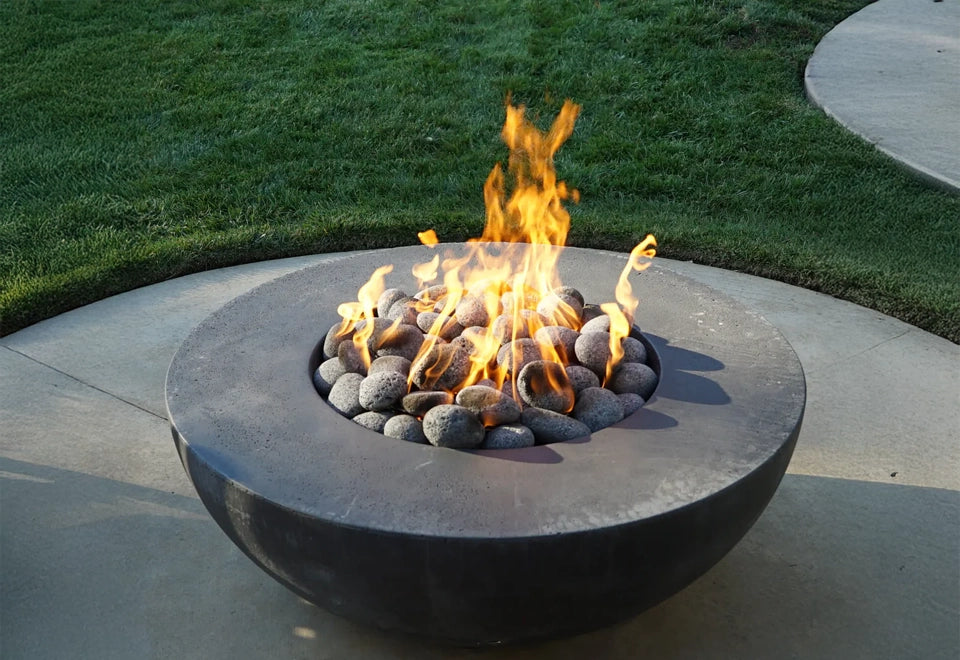 Modern round fire pit with natural lava rocks and vibrant flames, set on a concrete patio with lush green lawn in the background for an inviting outdoor space