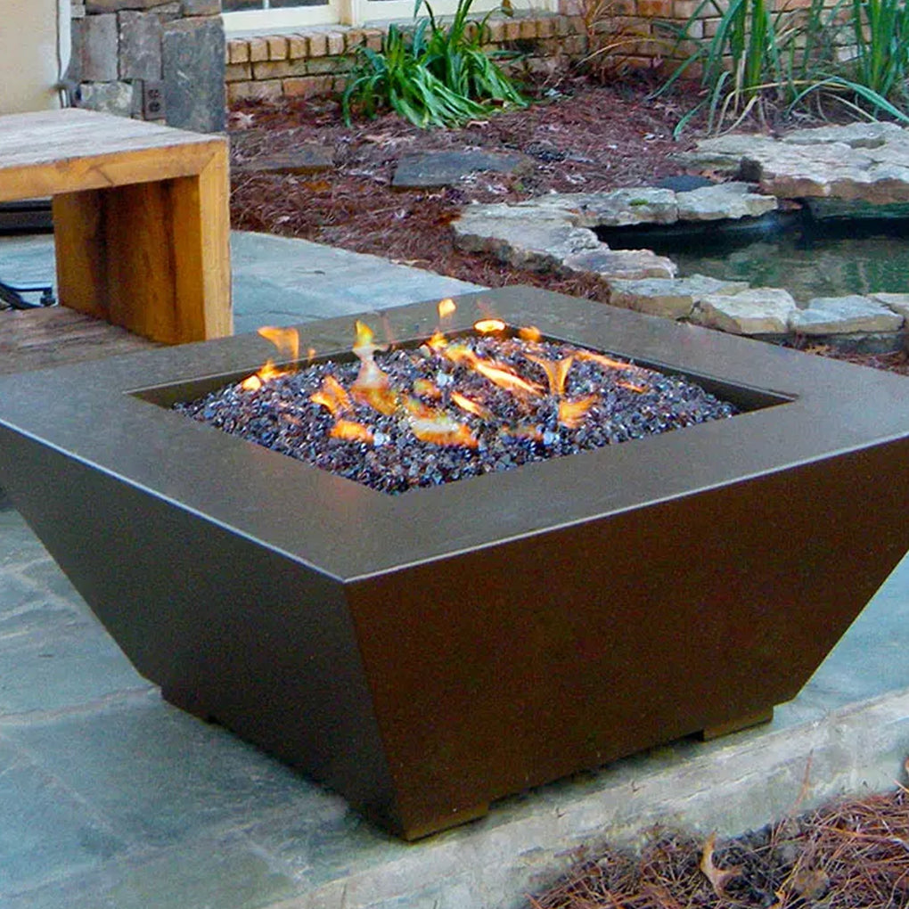 Square fire pit with dark-colored fire glass on a stone patio, set near a garden pond, creating a rustic outdoor ambiance