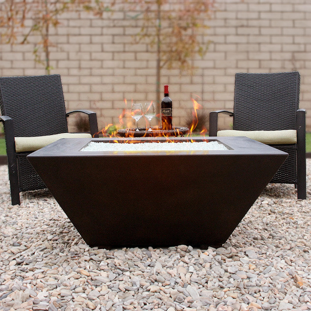 Modern trapezoidal fire pit with clear fire glass, surrounded by wicker chairs in an outdoor setting, featuring a bottle of wine and glasses