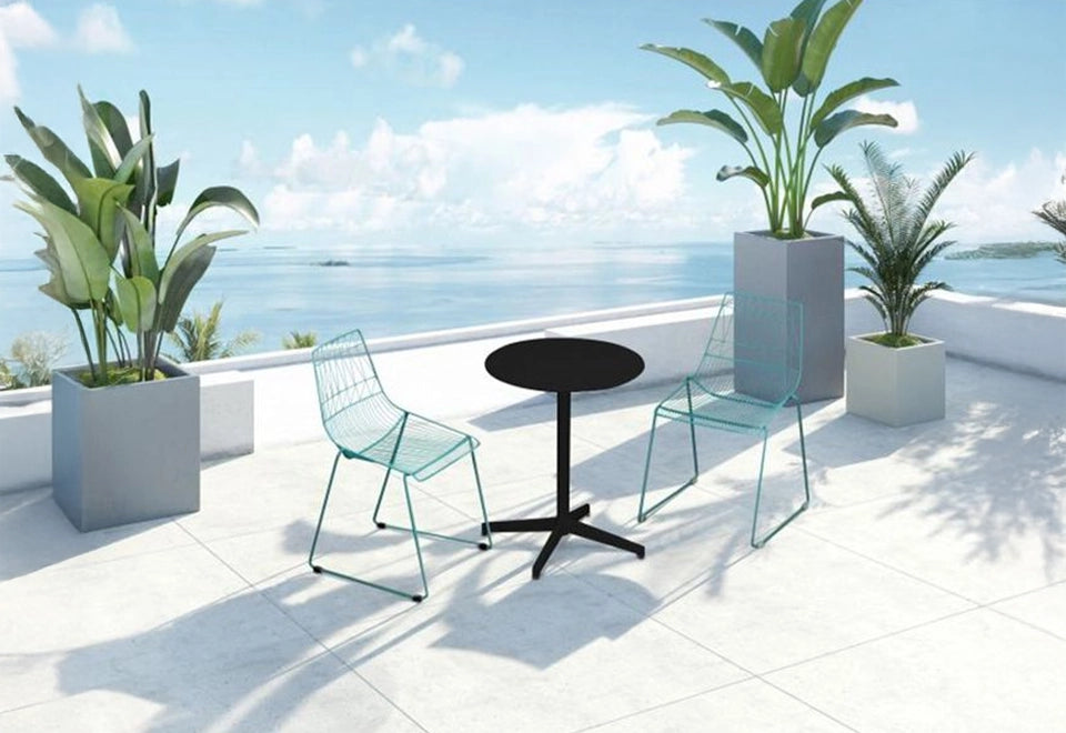 Contemporary balcony bistro set featuring teal wire chairs and a sleek black round table, surrounded by modern planters with tropical greenery and an ocean view.