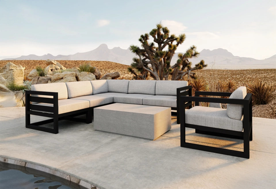 Modern patio sectional with a sleek black aluminum frame and light gray cushions, set in a desert oasis landscape with natural rock formations and Joshua trees.