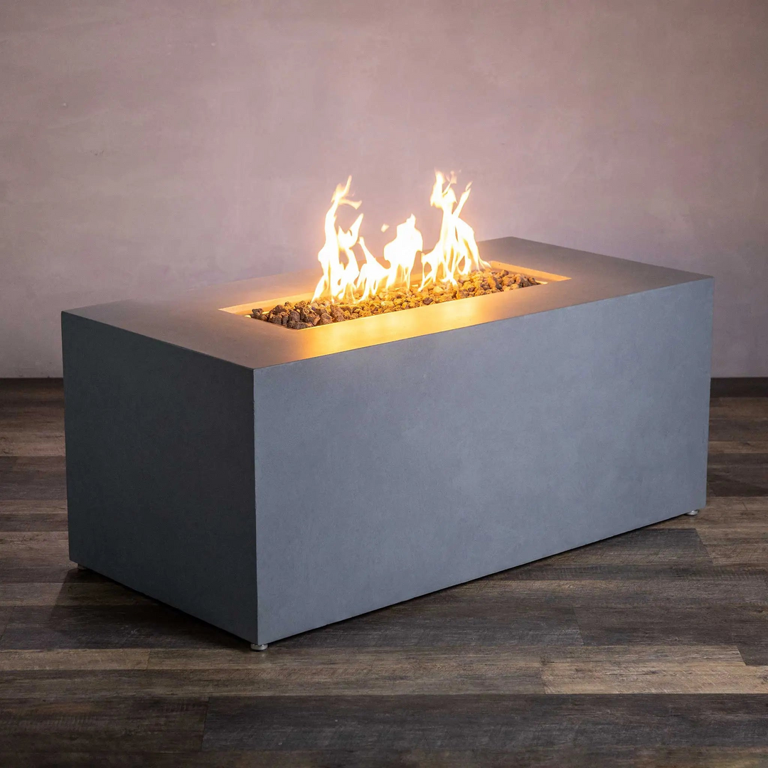 Modern rectangular fire pit with a sleek, concrete finish, featuring flames rising from a bed of lava rocks, placed indoors on a wooden floor against a plain, neutral wall.