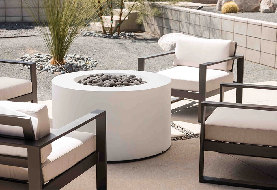 Stylish round white concrete fire pit with black lava rocks, enhancing a modern outdoor patio setting with comfortable lounge chairs.