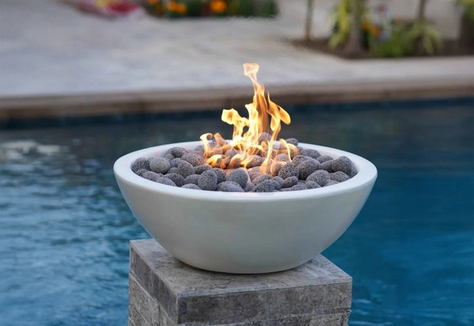 Modern white round gas fire bowl with smooth concrete finish and natural lava rock media, elegantly placed on a stone pedestal by the poolside. The vibrant flames create a warm and inviting ambiance, perfect for enhancing outdoor patio spaces and luxurious backyard fire pit installations.