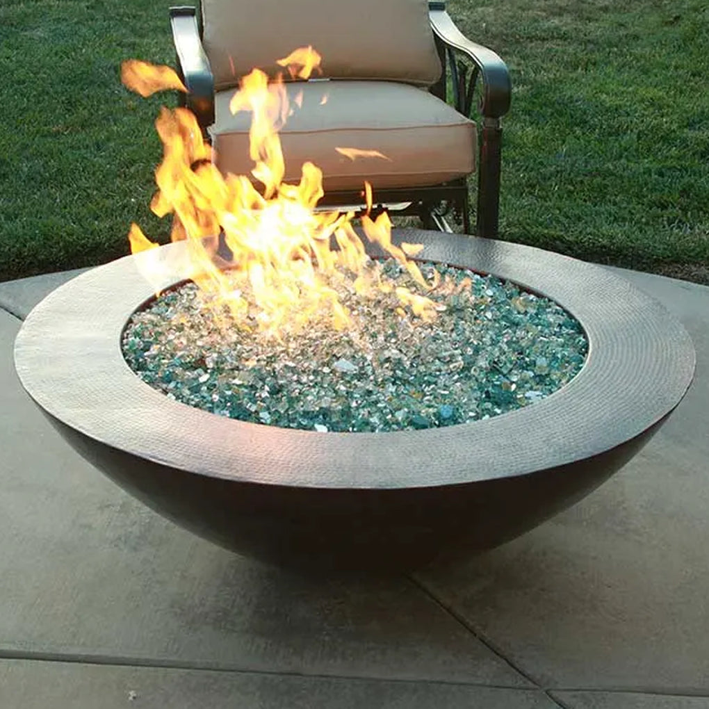 Round bowl-shaped fire pit with a wide rim, filled with reflective glass fire media, creating a stunning visual display of flames in an outdoor setting