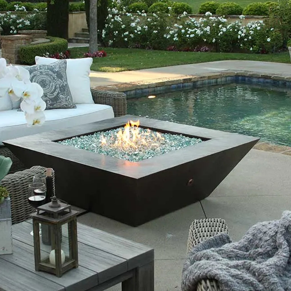 Square, angular fire pit filled with reflective glass fire media in a luxurious outdoor setting, situated poolside with comfortable seating and a well-maintained garden backdrop