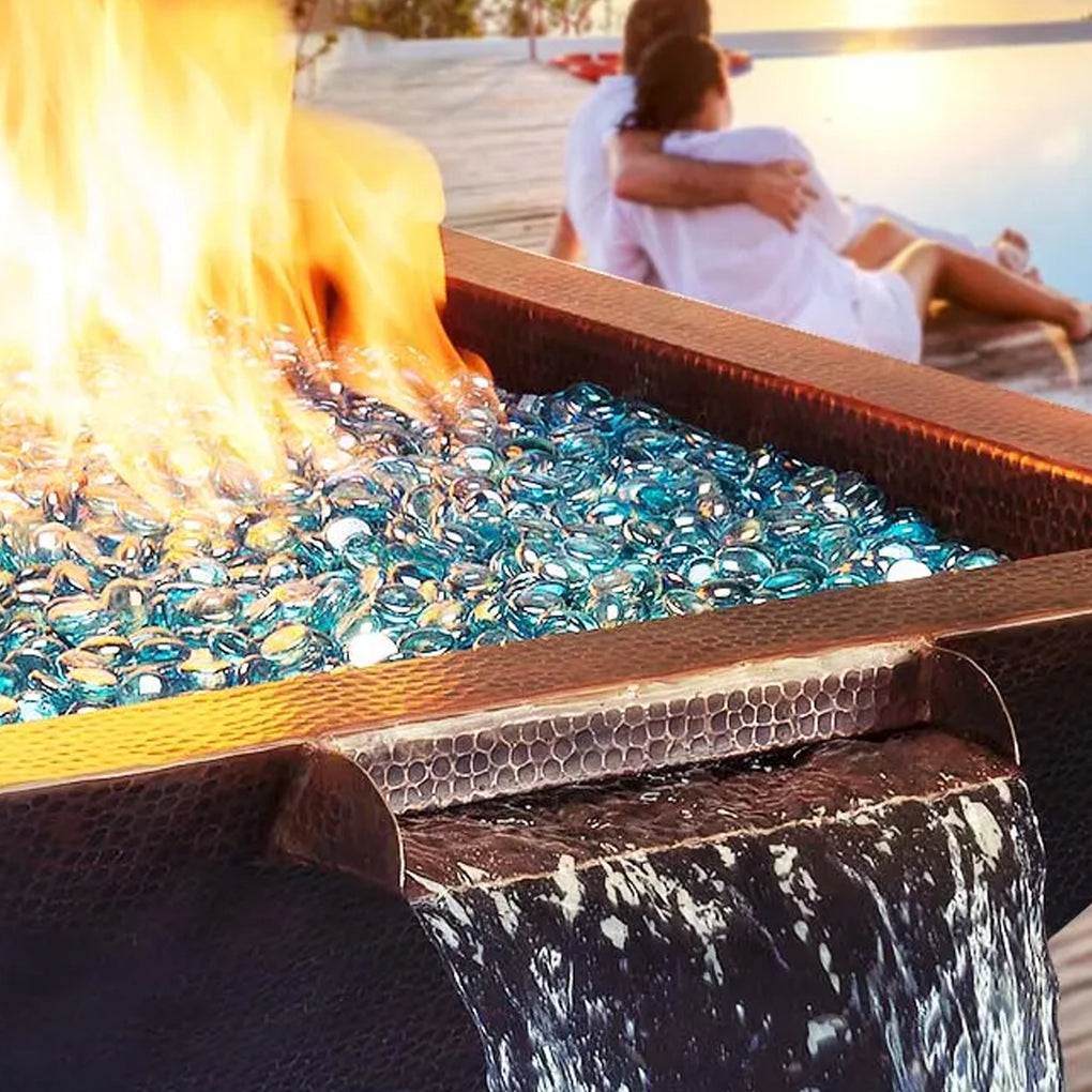 Close-up of a fire pit with blue glass fire media and a cascading waterfall feature, set against a romantic background of a couple sitting by the water.