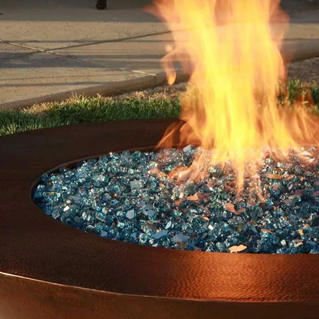 Close-up of a fire pit with blue fire glass media and vibrant flames rising, set against a background of pavement and grass