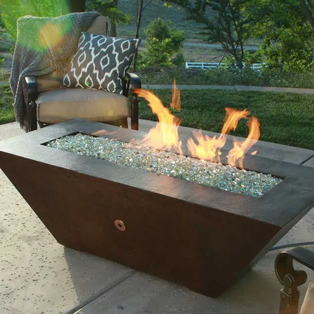 Modern rectangular fire pit filled with clear fire glass and vibrant flames in an outdoor setting with an armchair, blanket, and grassy background