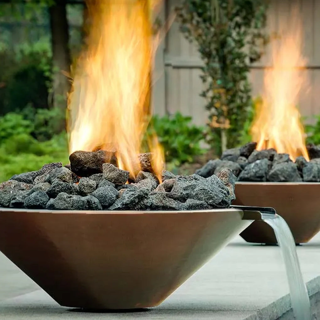 Modern fire bowls filled with dark lava rocks and cascading waterfalls in a serene outdoor setting with lush greenery and a wooden fence