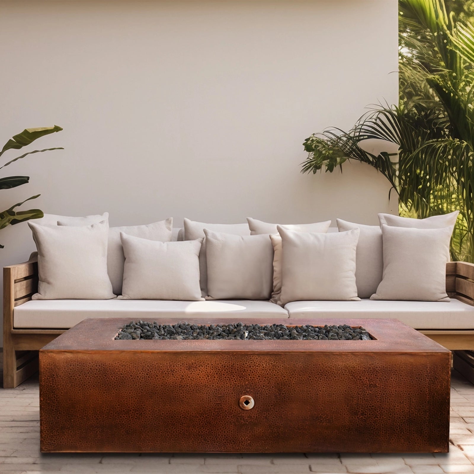 Modern outdoor space featuring a large copper fire pit table with black lava rocks, positioned in front of a white cushioned sofa, surrounded by tropical greenery