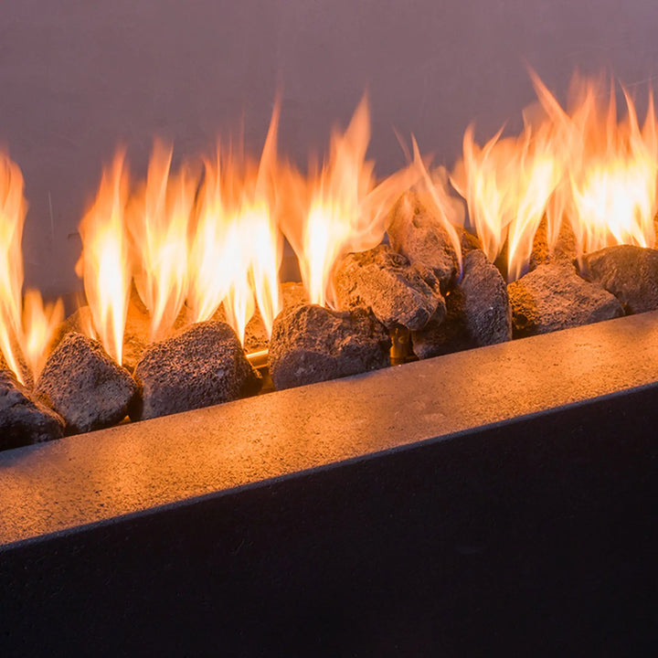 A close-up of large outdoor volcanic lava rocks with bright flames rising between them. The detailed texture of the porous volcanic stones contrasts beautifully with the soft glow of the fire, creating an inviting atmosphere. These large volcanic rocks are perfect for outdoor fire pits, offering both functionality and aesthetic appeal. Their rough, natural finish complements a variety of outdoor settings, adding warmth and a rugged, earthy look to fire features.
