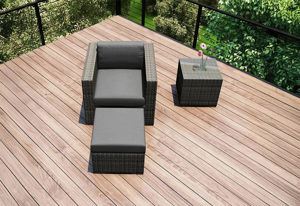 Contemporary outdoor wicker lounge chair with matching ottoman and side table, featuring dark gray cushions, set on a spacious wooden deck with lush greenery in the background.