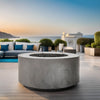 Prism Hardscapes Cilindro Gas Fire Pit showcased on a luxurious modern patio with a stunning ocean view in the background. This sleek, round gas fire pit in natural gray concrete complements the elegant outdoor seating area with plush cushions and neatly arranged plants, creating a serene and stylish atmosphere. Ideal for outdoor spaces like patios, gardens, and balconies, this natural gas fire pit offers both warmth and aesthetic appeal for relaxing evenings outdoors. 