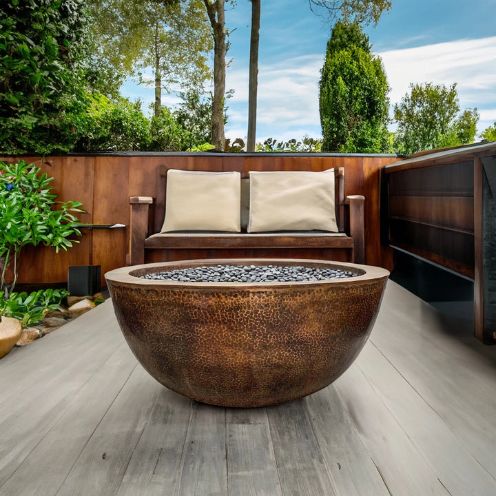 Prism Hardscapes 39" Edel Hospitality Moderno 1 Copper Fire Bowl, featured in a luxurious backyard setting with natural surroundings. This elegant copper fire pit is designed for outdoor use, making it a perfect centerpiece for any patio or garden space. The hammered copper finish offers a timeless and stylish touch. 