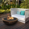 The Prism Hardscapes 48" Edel Hospitality Moderno 4 Gas Fire Pit displayed in a luxurious outdoor seating area surrounded by greenery and contemporary furniture. The hammered copper fire bowl beautifully complements the modern patio setup, creating an inviting and cozy atmosphere. This fire bowl adds a touch of warmth and sophistication to any outdoor space.