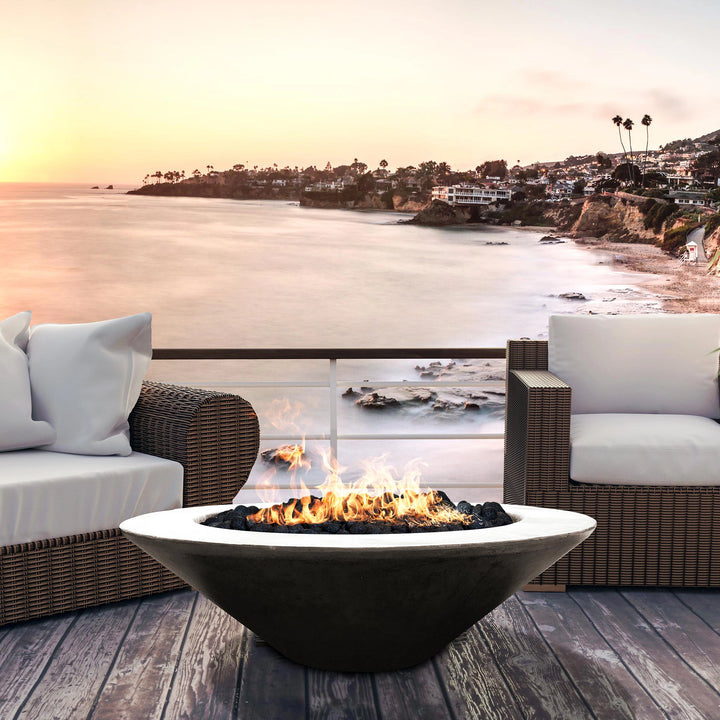 The Prism Hardscapes Embarcadero Gas Fire Pit in a natural gray finish set against a stunning coastal sunset, offering a serene ambiance for beachfront living. The wide-rimmed modern design seamlessly integrates with the wicker patio furniture, making it a perfect choice for luxurious seaside terraces. The gas fire pit's open flame adds warmth and a focal point for cozy evenings.