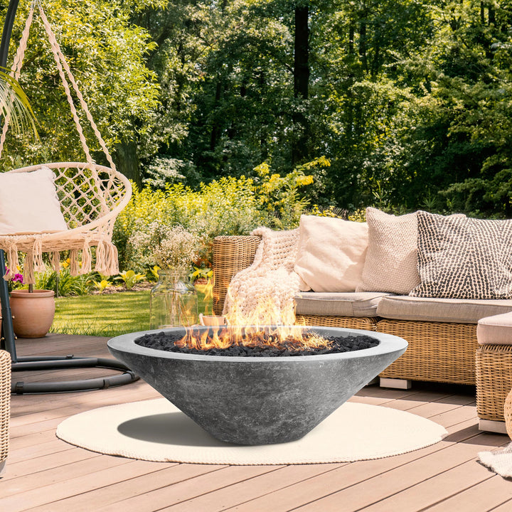 The Prism Hardscapes Embarcadero Gas Fire Pit in pewter, elegantly placed in a cozy garden setting surrounded by wicker furniture and lush greenery. The fire pit’s wide rim and industrial design complement the rustic charm of the space, creating a relaxing outdoor retreat with a warm, inviting ambiance. Perfect for backyard gatherings. 