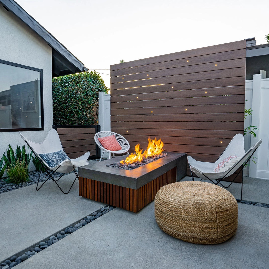 A stylish outdoor seating area featuring the Prism Hardscapes Fairmont Fire Table. This concrete fire pit table with a wooden slatted base is perfect for a cozy evening around the fire. The space is framed by a wood privacy wall, adding to the modern ambiance. The rectangular fire table offers warmth and design appeal for any small backyard or patio fire pit setup