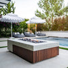 Prism Hardscapes Fairmont Fire Table elegantly placed by a pool with white umbrellas and modern lounge seating. The concrete fire table features a wooden slatted base, complemented by smooth river rocks. Ideal for poolside gatherings, this fire table adds a touch of sophistication to any outdoor patio space. Its sleek design creates an inviting backyard fire pit area.