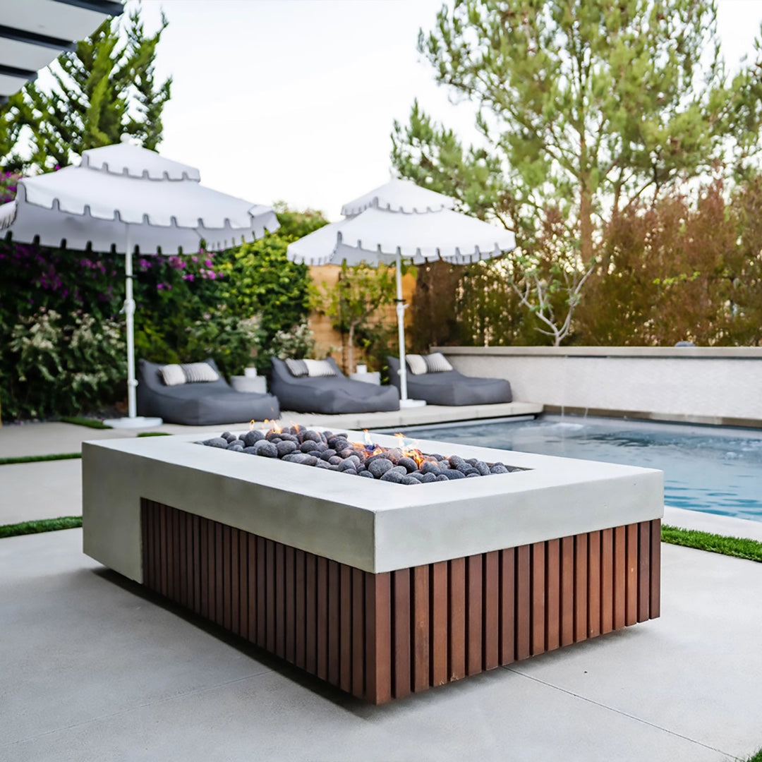 Prism Hardscapes Fairmont Fire Table elegantly placed by a pool with white umbrellas and modern lounge seating. The concrete fire table features a wooden slatted base, complemented by smooth river rocks. Ideal for poolside gatherings, this fire table adds a touch of sophistication to any outdoor patio space. Its sleek design creates an inviting backyard fire pit area.