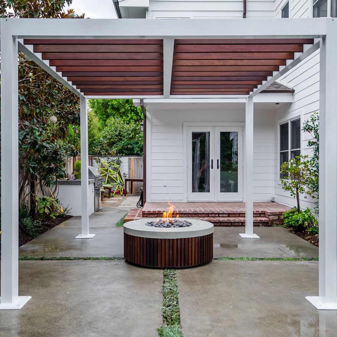 The Prism Hardscapes Hampton Fire Bowl is showcased in a sleek backyard under a modern pergola, adding warmth and style to the space. The round fire bowl, featuring a concrete top with vertical wooden panels, serves as the centerpiece of this chic outdoor patio. Perfect for backyard fire pits, modern outdoor entertaining areas, and creating a cozy fire pit atmosphere under a pergola. A perfect choice for a gas fire pit for stylish outdoor spaces.