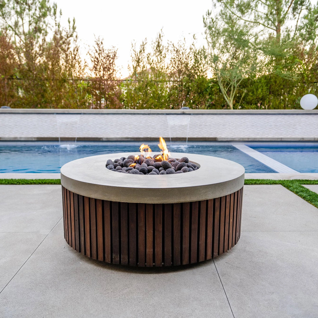 The Prism Hardscapes Hampton Fire Bowl sits next to a serene pool in a contemporary outdoor setting, featuring a round design with a smooth concrete top and warm wooden slatted panels. The fire pit creates a perfect focal point for backyard gatherings around the fire, combining modern design and function. Ideal for modern fire pits, luxury outdoor fire bowls, backyard poolside gatherings, and large fire pits for outdoor entertaining