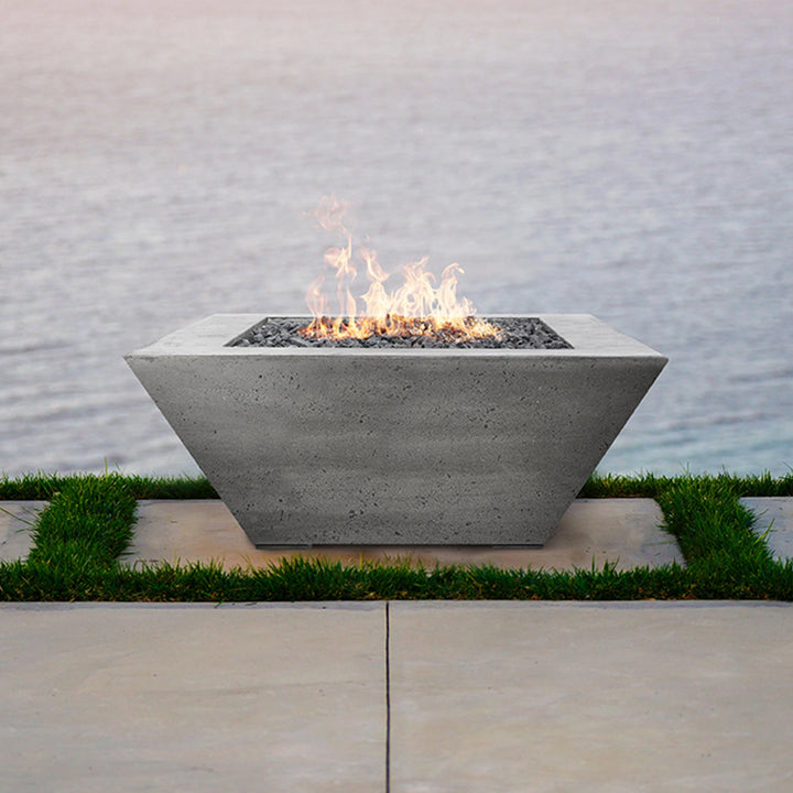 The Prism Hardscapes Lombard Gas Fire Pit in ebony finish is featured in a stylish patio setting, blending modern design with functionality. The fire pit's square, concrete form and minimalist design complement the outdoor lounge area, creating a perfect gathering spot. Whether for entertaining or relaxing by the fire, this gas fire pit delivers warmth and elegance to any outdoor space. It is ideal for use on patios or outdoor living areas with its sleek, architectural look.