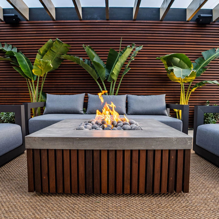 Prism Hardscapes Manchester Fire Table on a poolside patio, surrounded by wicker furniture and a tranquil setting. The fire table features a concrete top with wood panel detailing, offering a seamless blend of modern aesthetics and warmth. Ideal for backyard fire pit settings, outdoor gas fire bowls, and patio fire pits, adding both elegance and comfort to your outdoor living space.