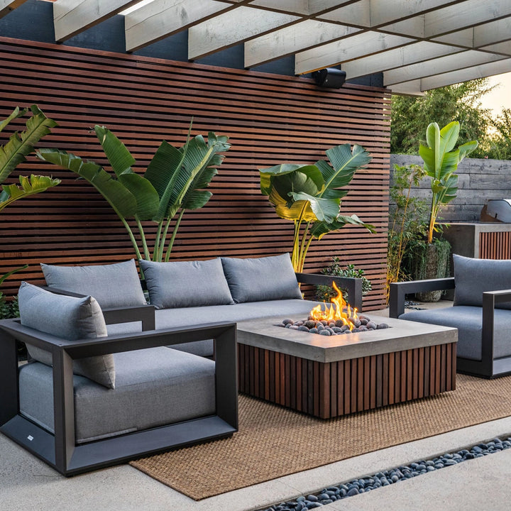 Prism Hardscapes Manchester Fire Table set in a stylish modern deck setting, featuring a rectangular concrete top with wooden paneling below. Surrounded by gray outdoor furniture, the fire table enhances the contemporary outdoor space. Ideal for backyard fire pit areas, gas fire bowls, and outdoor patio fire pits, this design brings a sleek and warm ambiance to your patio.