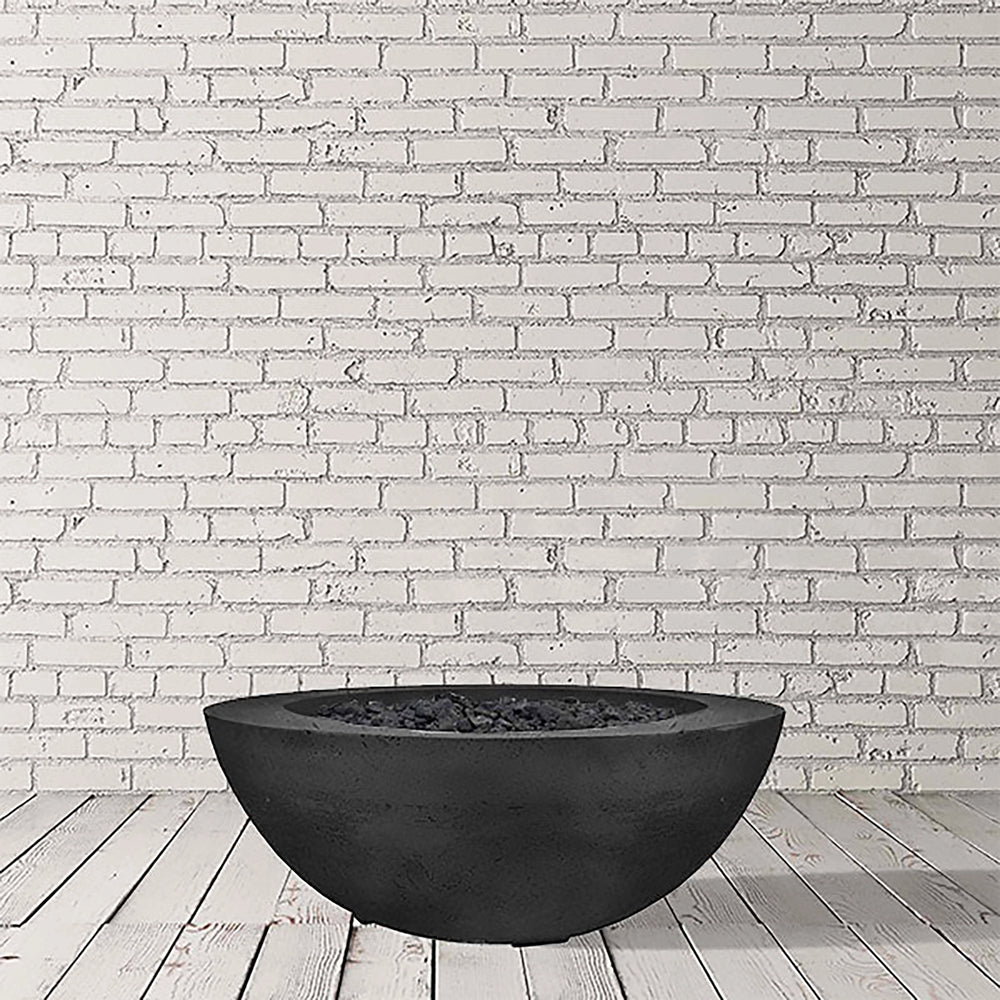 Prism Hardscapes Moderno 6 Gas Fire Bowl in an ebony black finish, photographed in a studio with a light brick wall. This sleek and contemporary gas fire pit is crafted from high-quality concrete, making it a modern fire pit perfect for commercial and outdoor residential spaces.