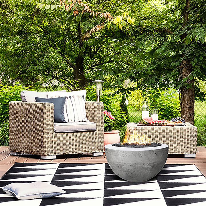 Prism Hardscapes Moderno 3 Gas Fire Bowl in a garden lounge setting, surrounded by wicker furniture and lush greenery. The fire pit, with its sleek concrete finish in a natural gray tone, adds a warm and inviting ambiance to this outdoor space. Ideal for a stylish backyard fire pit or patio centerpiece, the fire bowl enhances modern and rustic outdoor settings alike.