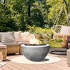 Prism Hardscapes Moderno 3 Gas Fire Bowl featured on a patio with cozy wicker furniture and a hammock chair. The fire pit, in a smooth natural gray concrete finish, creates an inviting focal point for outdoor gatherings. Surrounded by lush greenery, this fire bowl is ideal for outdoor spaces looking for a blend of comfort and style. Perfect for adding warmth to patios, decks, or backyards. 