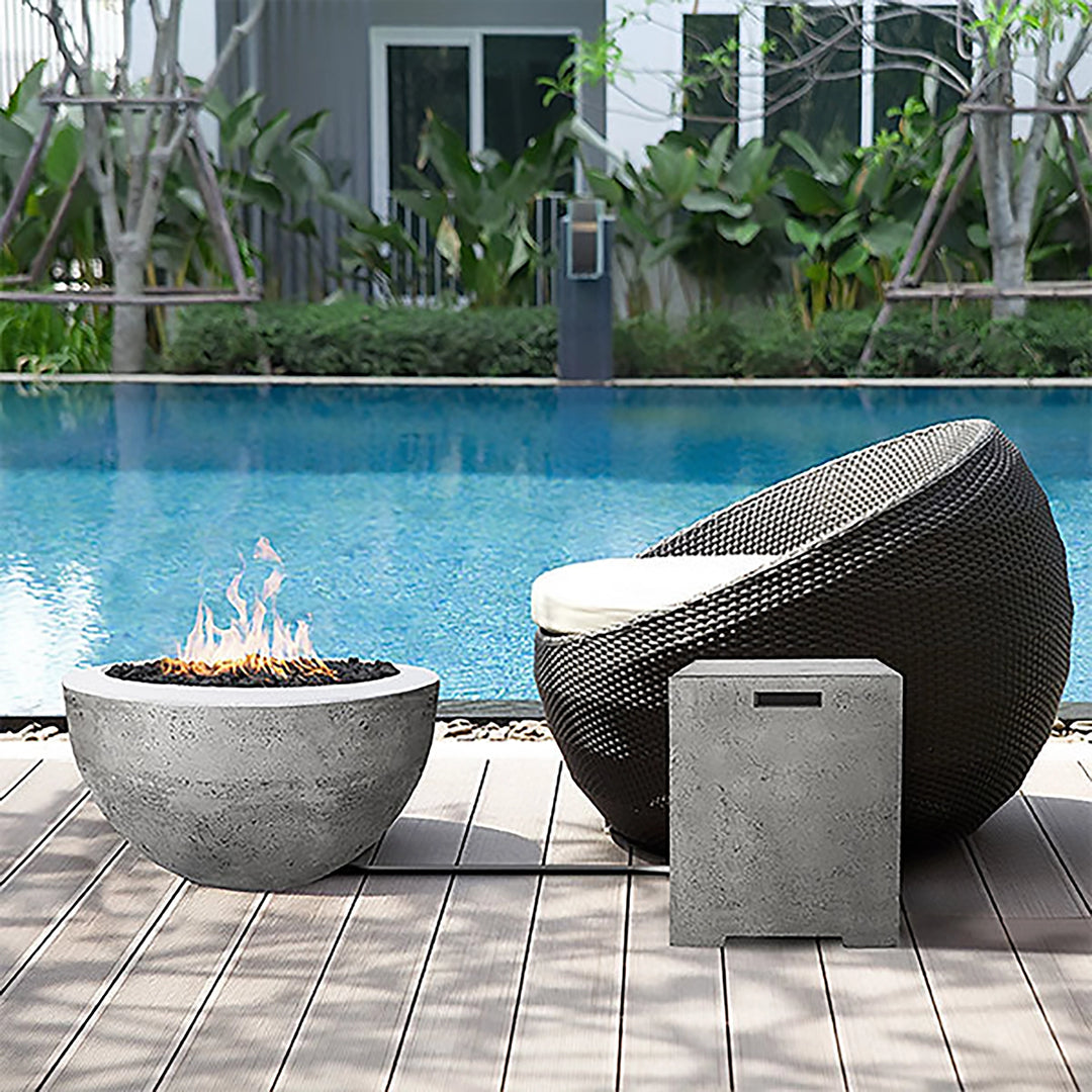 Prism Hardscapes Moderno 3 Gas Fire Bowl placed poolside, creating a serene atmosphere with its smooth concrete design in a natural gray finish. The fire pit sits next to a modern, circular outdoor chair, offering an ideal spot for relaxation by the pool. Perfect for enhancing outdoor spaces with its modern aesthetics, this gas fire pit is an excellent choice for a luxury backyard, patio, or poolside setting. 