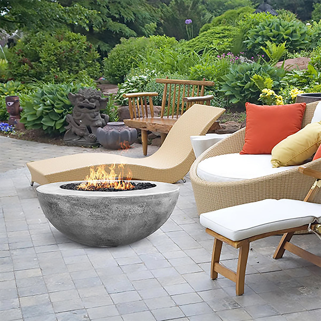 A beautiful backyard lounge featuring the Prism Hardscapes Moderno 5 Gas Fire Bowl in natural gray, blending seamlessly into the lush garden setting. With its sleek, modern design, the fire pit stands out as a functional and elegant centerpiece for a relaxing outdoor patio space. The flames flicker above the lava rocks, making it a perfect addition to this luxurious backyard retreat. 