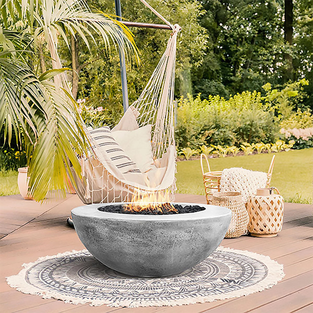 This image highlights the Prism Hardscapes Moderno 5 Gas Fire Bowl in pewter, placed on a cozy deck next to a relaxing hammock under tropical greenery. The fire pit emits gentle flames, creating a warm and serene outdoor space perfect for unwinding. The mix of the woven hammock and the sleek design of the fire pit offers an inviting contrast, enhancing both modern and bohemian vibes.