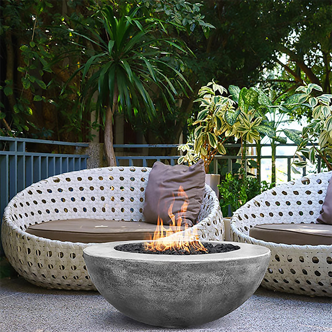 The Prism Hardscapes Moderno 5 Gas Fire Bowl in a sophisticated pewter finish is featured in a luxury outdoor lounge, surrounded by lush greenery and modern seating. Flames rise from black lava rocks, providing a warm and stylish focal point for the outdoor space. Ideal for contemporary outdoor designs, this fire pit enhances both comfort and aesthetic appeal.