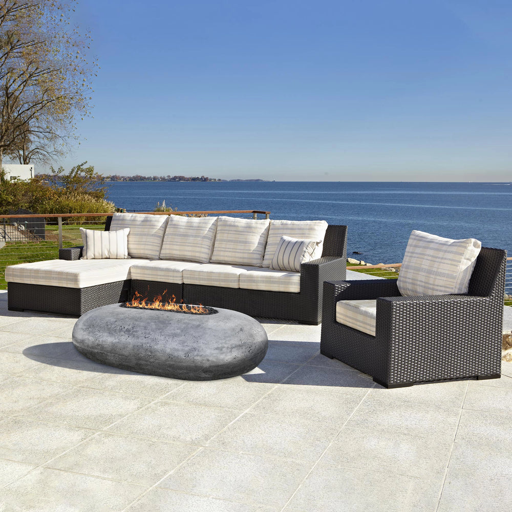 Prism Hardscapes Pebble Gas Fire Pit in a modern seaside outdoor setting, featuring a luxurious sofa set and a scenic view of the ocean. This natural gas fire pit offers a sleek, smooth pebble design, perfect for enhancing the aesthetics of any patio or outdoor living space. Ideal for backyard gatherings and designed with weather-resistant materials, this large fire pit blends seamlessly into modern outdoor furniture. Outdoor gas firepit, luxury fire pit, modern fire pit, backyard fire pit, outdoor living.