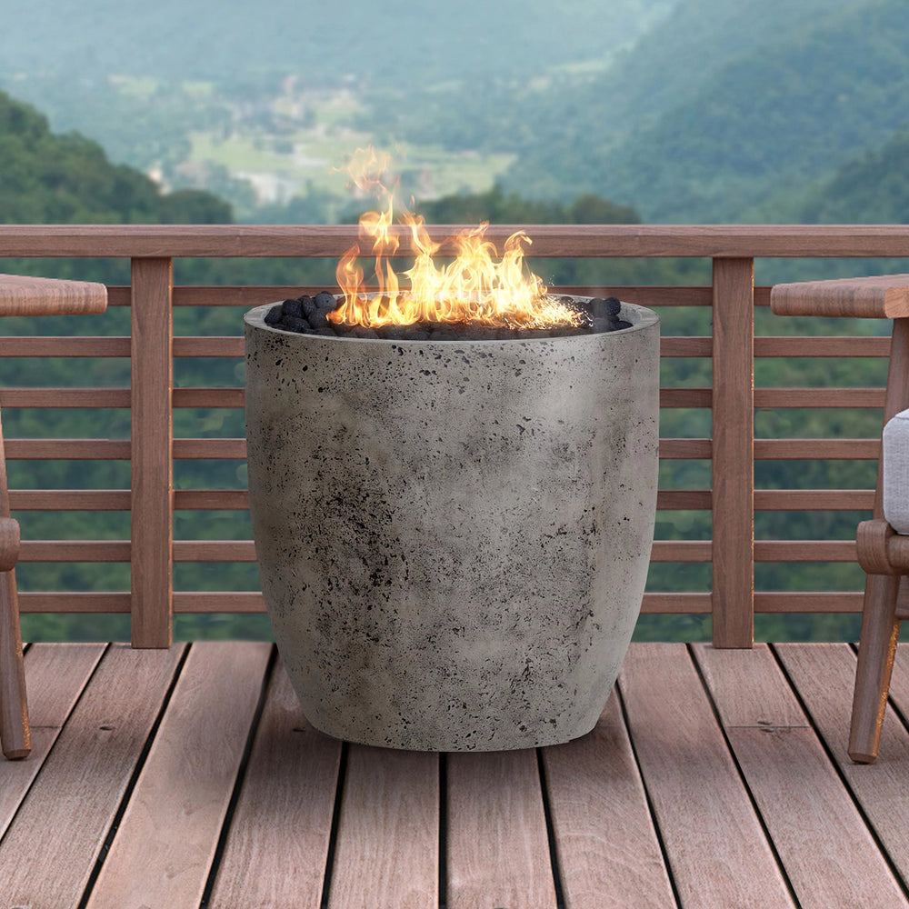 The Prism Hardscapes Pentola 2 Gas Fire Column in a natural textured finish, placed on a wooden deck with flames rising from the top. Perfect for outdoor patios or backyards, this fire bowl enhances the ambiance with its durable, rustic design. Ideal for modern outdoor living spaces. Keywords: fire pit, outdoor fire column, backyard fire pit, patio fire pit, natural gas fire column