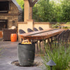 Prism Hardscapes Pentola 2 Gas Fire Column in a pewter finish, shown in an elegant patio setting beside a long wooden table and lush greenery. The sleek fire bowl adds both warmth and style to any outdoor space, creating a cozy gathering area for socializing. Keywords: patio fire pit, outdoor fire bowl, natural gas fire pit, backyard fire feature, pewter fire column.