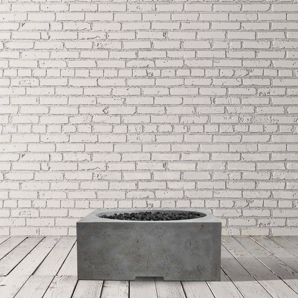 Studio shot of the Prism Hardscapes Piazza Gas Fire Pit in Pewter, placed against a white brick wall backdrop. This modern concrete fire pit features a durable gray finish, adding an industrial yet refined look to any outdoor setting. The low-profile design makes it ideal for patios, backyard fire pits, or commercial fire pit installations. Perfect for those seeking a contemporary fire pit with a natural gas option, this model blends functionality with aesthetic appeal.