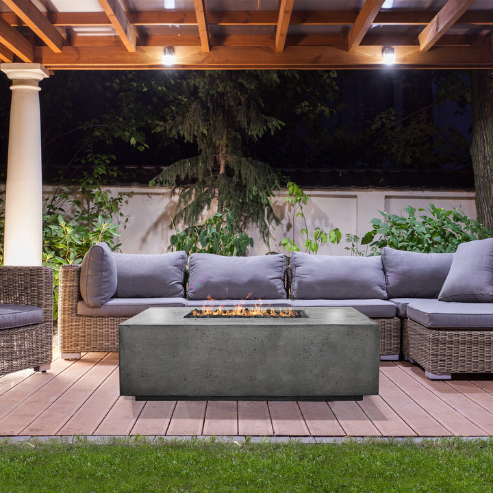 The Prism Hardscapes Portos 58 Propane Fire Table in a gray finish is shown in a stylish outdoor setting with modern seating, including patterned cushions and wicker chairs. The fire table features clean lines and a rectangular design, perfect for a contemporary patio. It is surrounded by a wood deck and black-and-white geometric rug, creating a warm ambiance for backyard gatherings. Ideal for outdoor fire pits, this large rectangular gas fire pit serves as a modern centerpiece for fire tables on the patio 